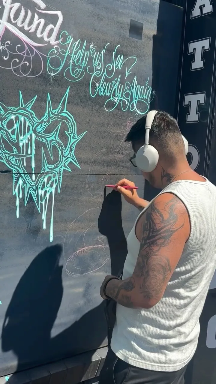 Artist drawing turquoise chalk lettering on the True Addiction shopfront, vertical TATTOO signage visible